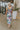 Back view of model wearing the In Bloom Midi Dress that has white fabric with a purple, blue, green and pink watercolor floral design, ruching, a v-neck, thick straps, a a side slit, and a lining