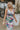 Close front view of model wearing the In Bloom Midi Dress that has white fabric with a purple, blue, green and pink watercolor floral design, ruching, a v-neck, thick straps, a a side slit, and a lining