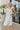 Front view of model wearing the Picture Perfect Midi Dress in White that has sheer white fabric with a checkered pattern, a white lining, a tiered midi-length body, side pockets, and tiered cap sleeves.