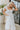 Front view of model wearing the Picture Perfect Midi Dress in White that has sheer white fabric with a checkered pattern, a white lining, a tiered midi-length body, side pockets, and tiered cap sleeves.