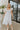 Front view of model wearing the Picture Perfect Midi Dress in White that has sheer white fabric with a checkered pattern, a white lining, a tiered midi-length body, side pockets, and tiered cap sleeves.