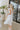 Back/side view of model wearing the Picture Perfect Midi Dress in White that has sheer white fabric with a checkered pattern, a white lining, a tiered midi-length body, side pockets, and tiered cap sleeves.