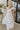 Frontal side view of model wearing the Picture Perfect Midi Dress in White that has sheer white fabric with a checkered pattern, a white lining, a tiered midi-length body, side pockets, and tiered cap sleeves.