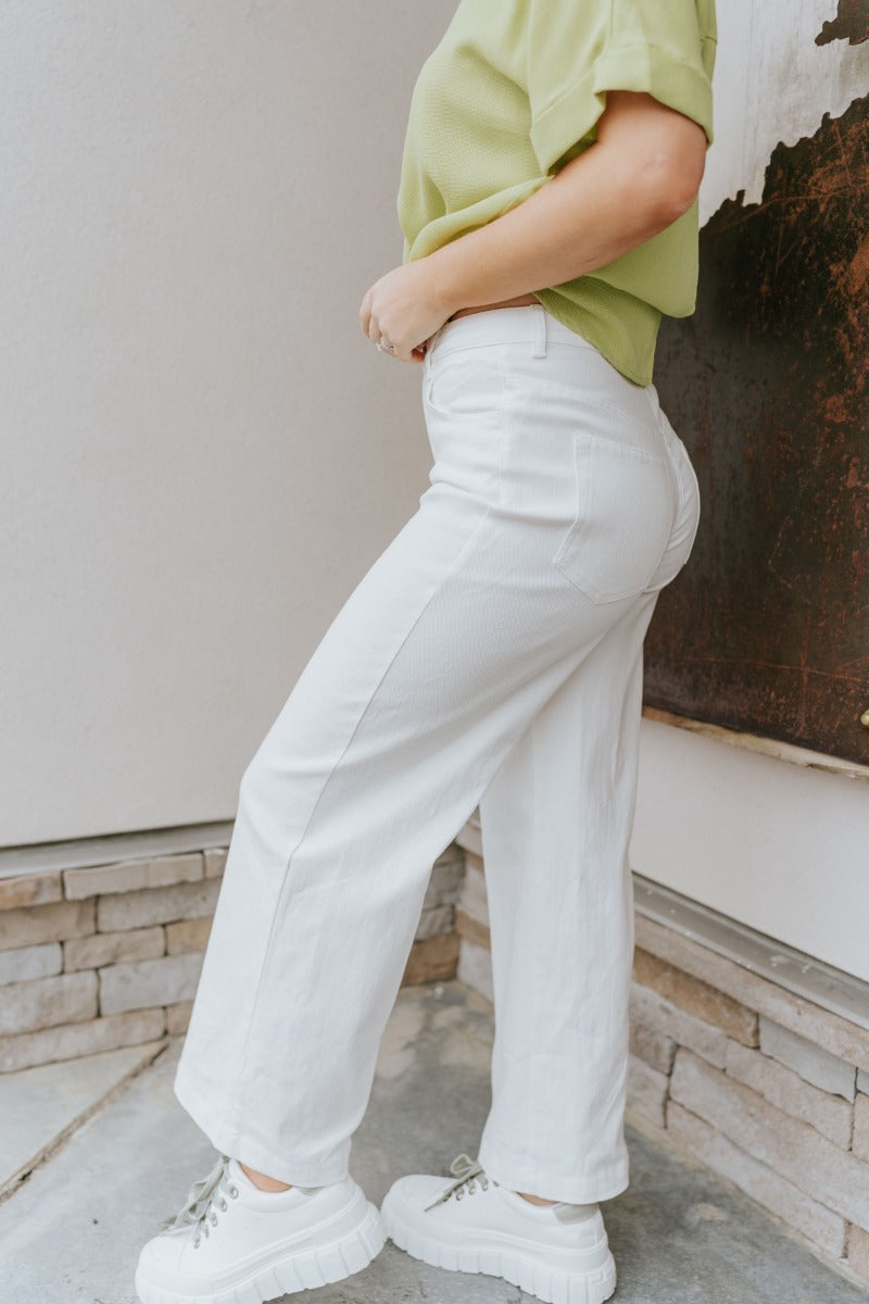 Side view of model wearing the Where Have You Been Pants which features white denim fabric, two front pockets, two back pockets, belt loops, a front button and zipper closure, and cropped flare legs.