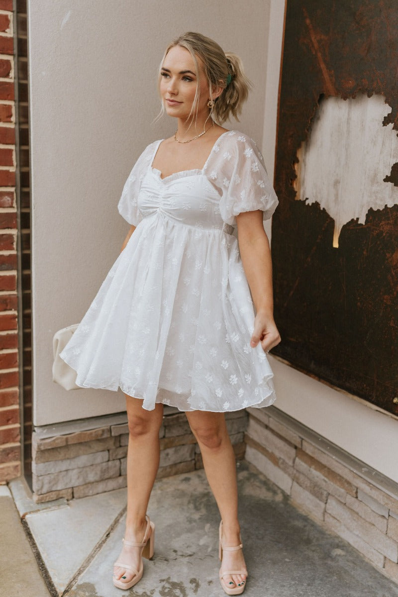 Full body front view of model wearing the Happily Ever After Dress that has white sheer fabric with a white floral embroidery, a mini-length hem, a square neck, ruched detailing, a smocked back, and short puff sleeves