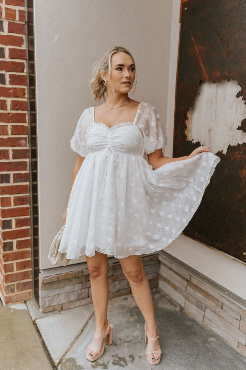 Full body front view of model wearing the Happily Ever After Dress that has white sheer fabric with a white floral embroidery, a mini-length hem, a square neck, ruched detailing, a smocked back, and short puff sleeves