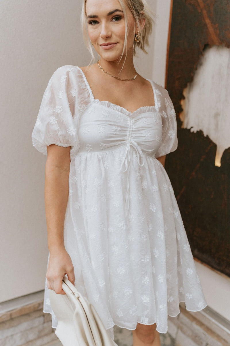 Front view of model wearing the Happily Ever After Dress that has white sheer fabric with a white floral embroidery, a mini-length hem, a square neck, ruched detailing, a smocked back, and short puff sleeves