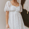 Front view of model wearing the Happily Ever After Dress that has white sheer fabric with a white floral embroidery, a mini-length hem, a square neck, ruched detailing, a smocked back, and short puff sleeves