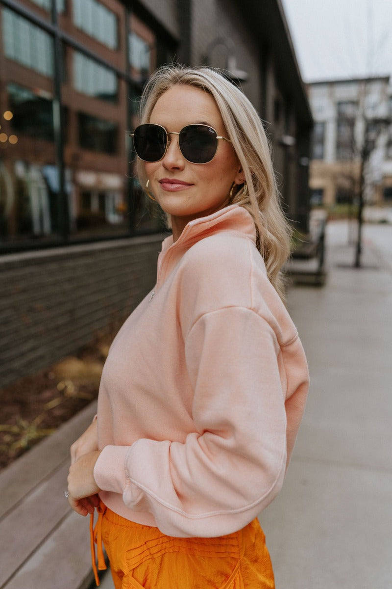 Side view of model wearing the Coastal Breeze Pullover that has peach colored fabric, a cropped waist, a monochromatic quarter zip-up, a high neckline, and long sleeves with cuffs