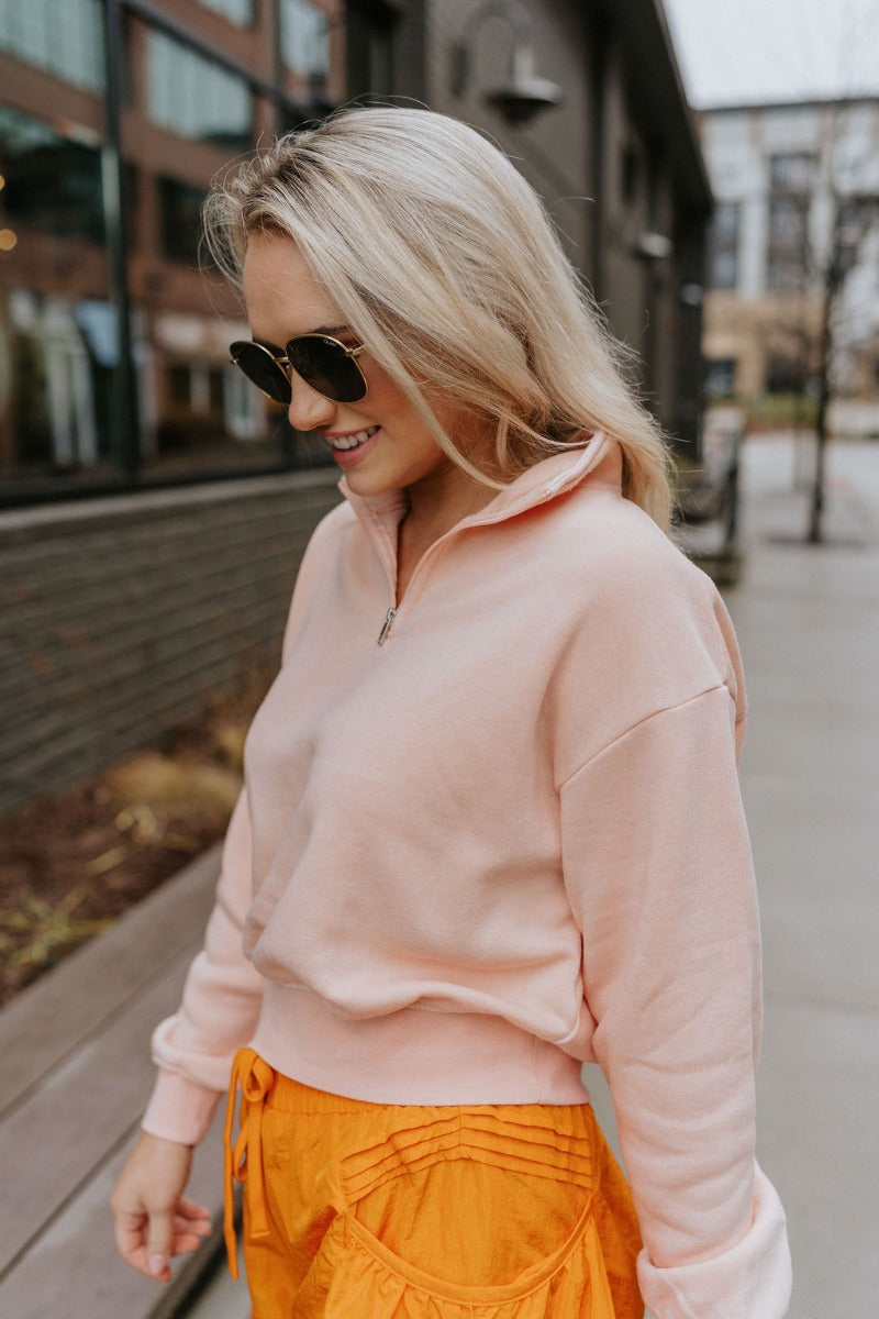 Frontal side view of model wearing the Coastal Breeze Pullover that has peach colored fabric, a cropped waist, a monochromatic quarter zip-up, a high neckline, and long sleeves with cuffs