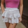 Front view of model wearing the It's Complicated Satin Skort in White which features white satin fabric, a tiered body with ruffle details, an elastic drawstring waist, and shorts lining.