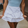 Front view of model wearing the Dare To Be Different Skort in White which features white denim fabric, a tiered skirt with shorts lining, brown contrast stitching, and an exposed back zipper.