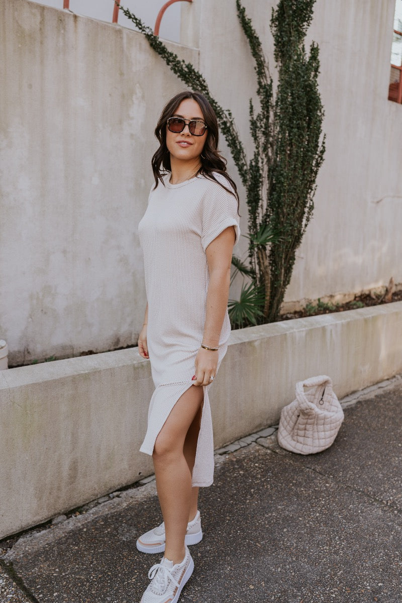 side view of model wearing The Cecile Ribbed Midi Dress features light beige and dark beige textured ribbing, a round neckline, short sleeves with cuffs, side slits, and a slight high-low hemline.