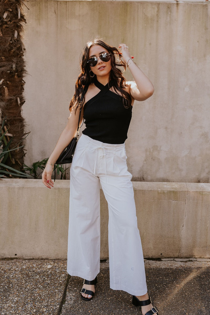 front view of model wearing The Isla White Pleated Pants features white knit fabric, wide pant legs, two side pockets, front zipper with hook closure and belt loops with tie around the waist.