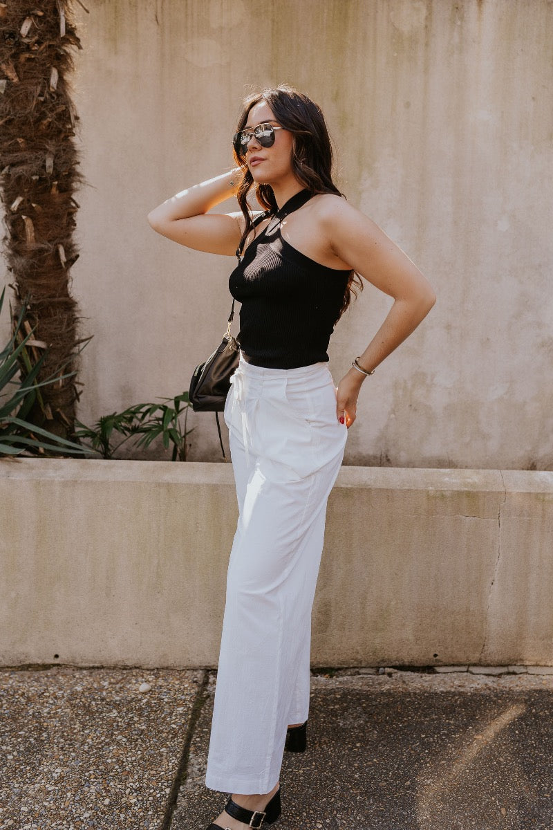 side view of model wearing The Isla White Pleated Pants features white knit fabric, wide pant legs, two side pockets, front zipper with hook closure and belt loops with tie around the waist.