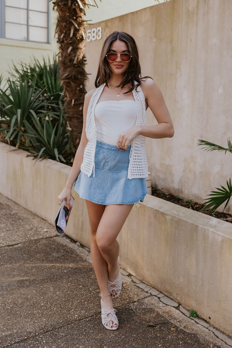 front view of model wearing The Azure Denim Ruffle Mini Skirt features washed light blue fabric, shorts lining, mini length, ruffle skirt overlay, flared skirt hem and side zipper closure.