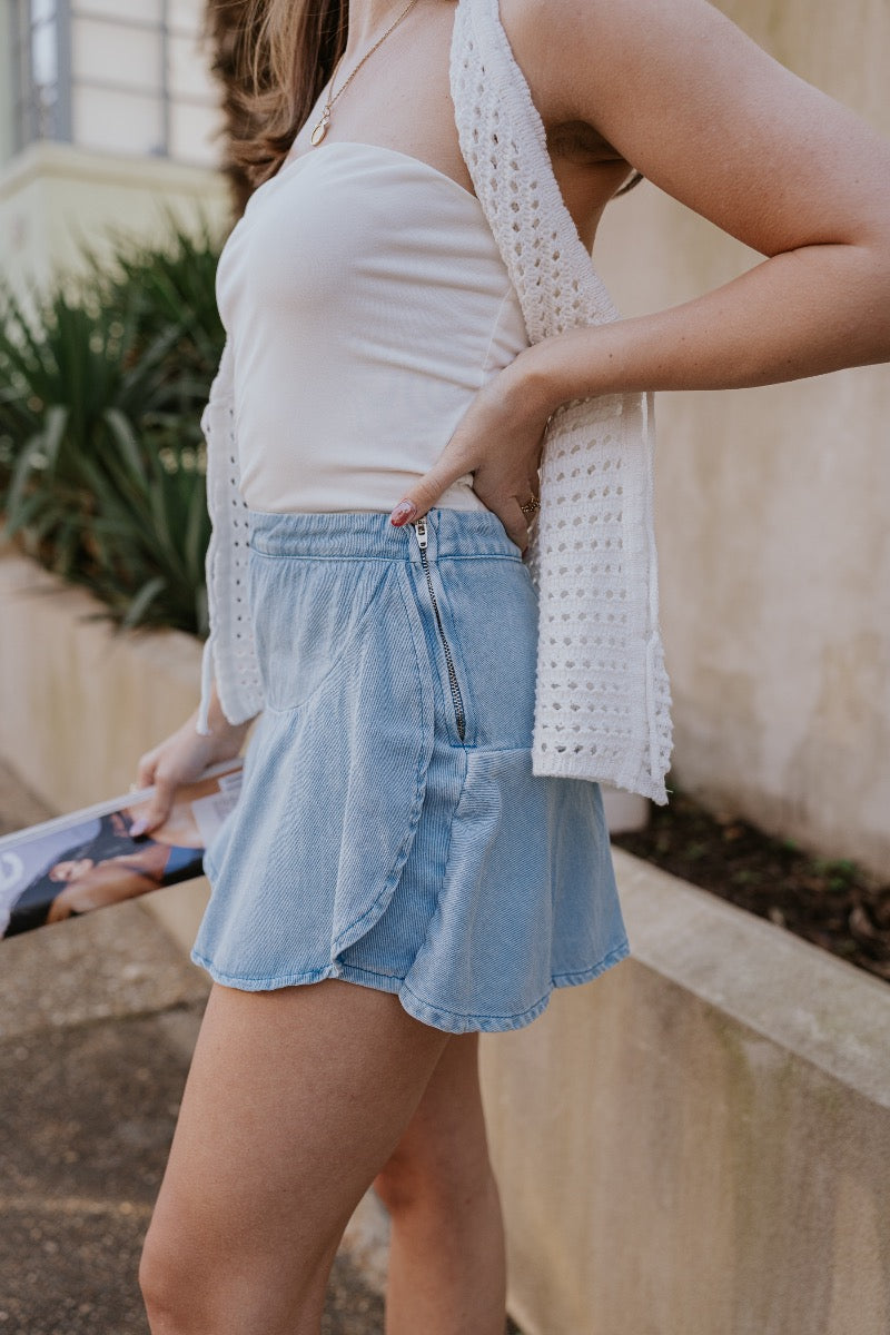 side detail view of model wearing The Azure Denim Ruffle Mini Skirt features washed light blue fabric, shorts lining, mini length, ruffle skirt overlay, flared skirt hem and side zipper closure.