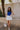 front view of model wearing The Azure White Denim Ruffle Mini Skirt features off white denim fabric, shorts lining, mini length, a ruffled skirt overlay, a flared skirt hem, and a side zipper closure.