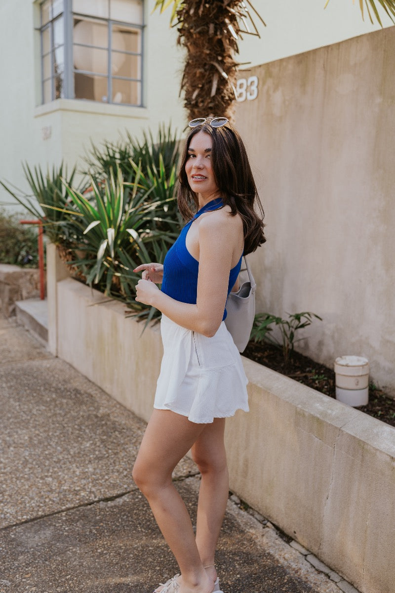 side view of model wearing The Azure White Denim Ruffle Mini Skirt features off white denim fabric, shorts lining, mini length, a ruffled skirt overlay, a flared skirt hem, and a side zipper closure.