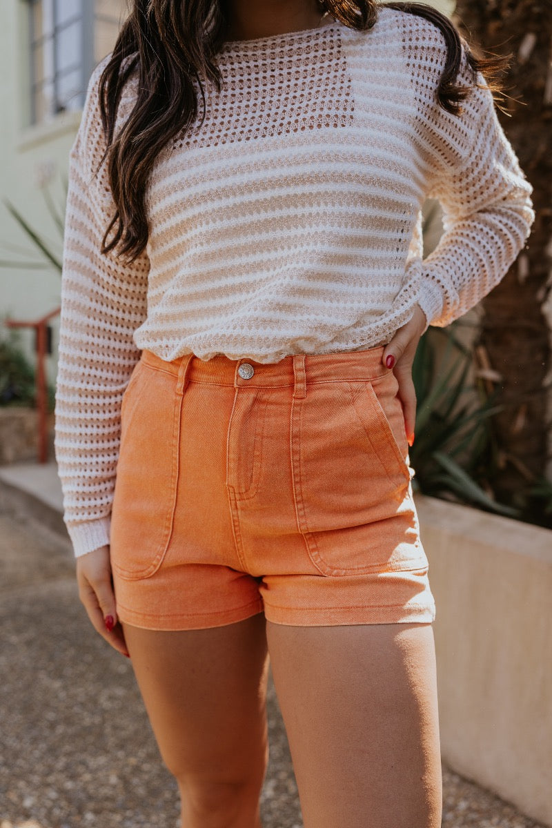 front view of model wearing The Monaco Orange Denim Shorts features washed orange denim fabric, two front pockets, two back pockets, front zipper with button closure and belt loops.