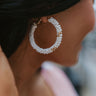 Close up view of model wearing the Enchanted Views Earring which features large closed hoops covered with white and gold beads.