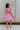 Back view of model wearing the Heart Breaker Dress in Pink that features pink fabric with a lining, a mini-length hem, a baby doll style, a v neckline, elastic straps with ruffles, and an open back with a bow detail.
