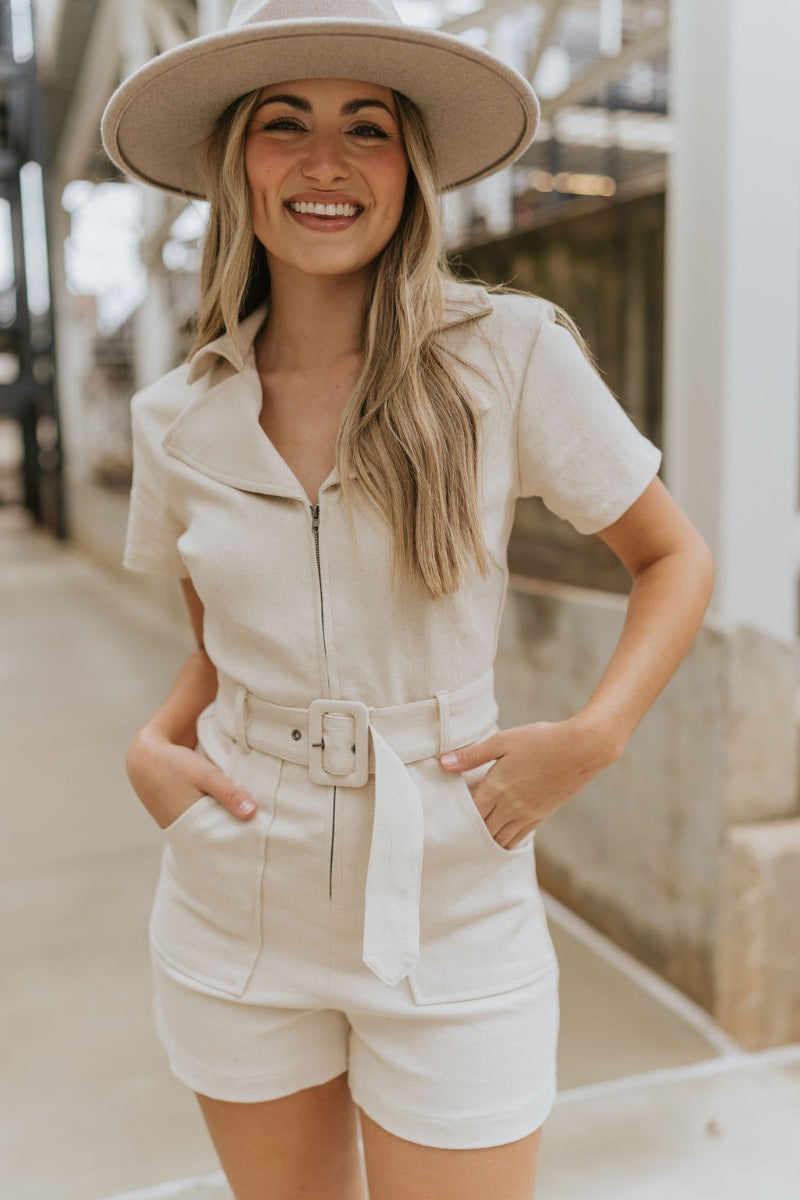 Front view of model wearing the Dakota Denim Romper in Oatmeal that has oatmeal denim fabric, front pockets, a front zipper, a collare, a belt at the waistline, and short sleeves