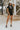 Full body front view of model wearing the Dakota Denim Romper in Black that has black denim fabric, front pockets, a hidden front zipper, a collared neckline, a monochromatic belt at the waistline, and short sleeves
