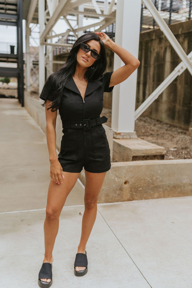 Full body front view of model wearing the Dakota Denim Romper in Black that has black denim fabric, front pockets, a hidden front zipper, a collared neckline, a monochromatic belt at the waistline, and short sleeves