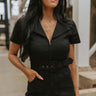 Front view of model wearing the Dakota Denim Romper in Black that has black denim fabric, front pockets, a hidden front zipper, a collared neckline, a monochromatic belt at the waistline, and short sleeves