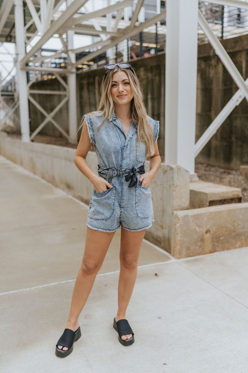 Full body front view of model wearing the Are You Ready Romper that has washed lightweight denim fabric, front pockets, two back pockets, a hidden front snap closure, a collar, short sleeves, and  frayed hems.