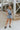 Full body front view of model wearing the Are You Ready Romper that has washed lightweight denim fabric, front pockets, two back pockets, a hidden front snap closure, a collar, short sleeves, and  frayed hems.