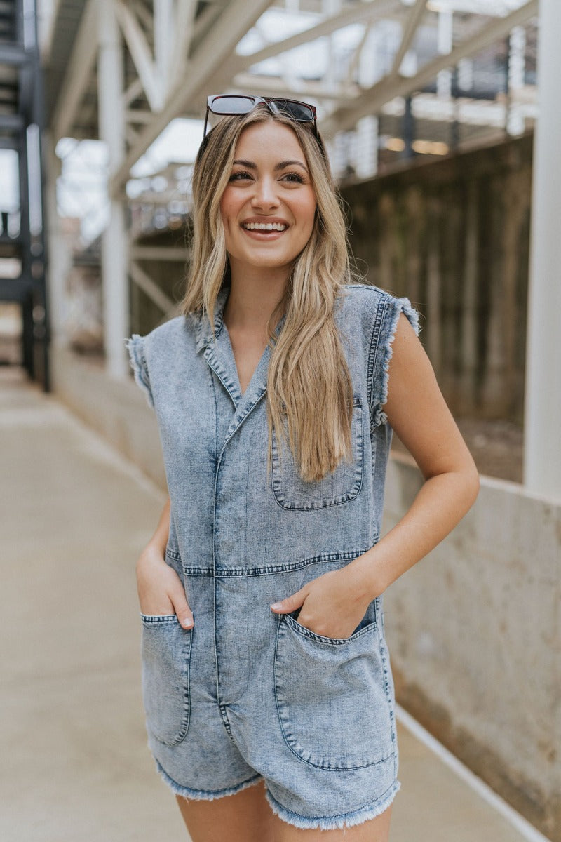 Front view of model wearing the Are You Ready Romper that has washed lightweight denim fabric, front pockets, two back pockets, a hidden front snap closure, a collar, short sleeves, and  frayed hems.