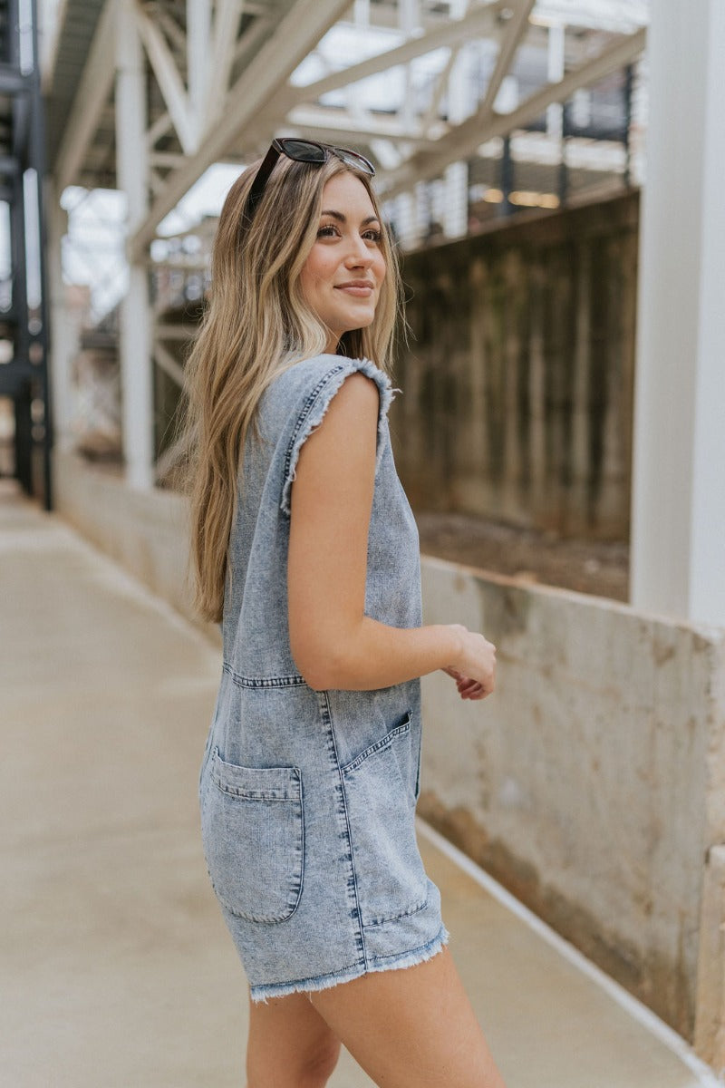 Side view of model wearing the Are You Ready Romper that has washed lightweight denim fabric, front pockets, two back pockets, a hidden front snap closure, a collar, short sleeves, and  frayed hems.