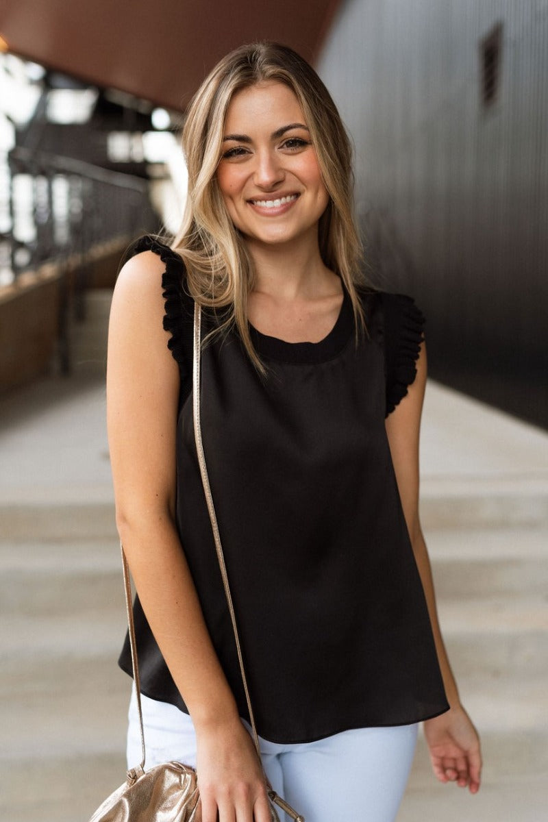 Front view of model wearing the Everything You Love Top in Black which features black satin fabric, a ribbed round neckline, and a sleeveless body with ruffled details.