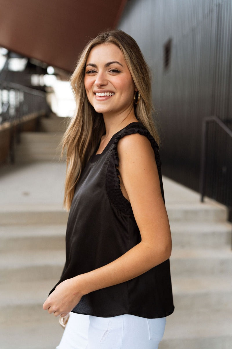 Side view of model wearing the Everything You Love Top in Black which features black satin fabric, a ribbed round neckline, and a sleeveless body with ruffled details.