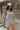 Side view of model wearing the Head In The Clouds Romper which features light grey knit fabric, pockets on each side, a quarter button-up with wooden buttons, a round neckline, and thick straps.