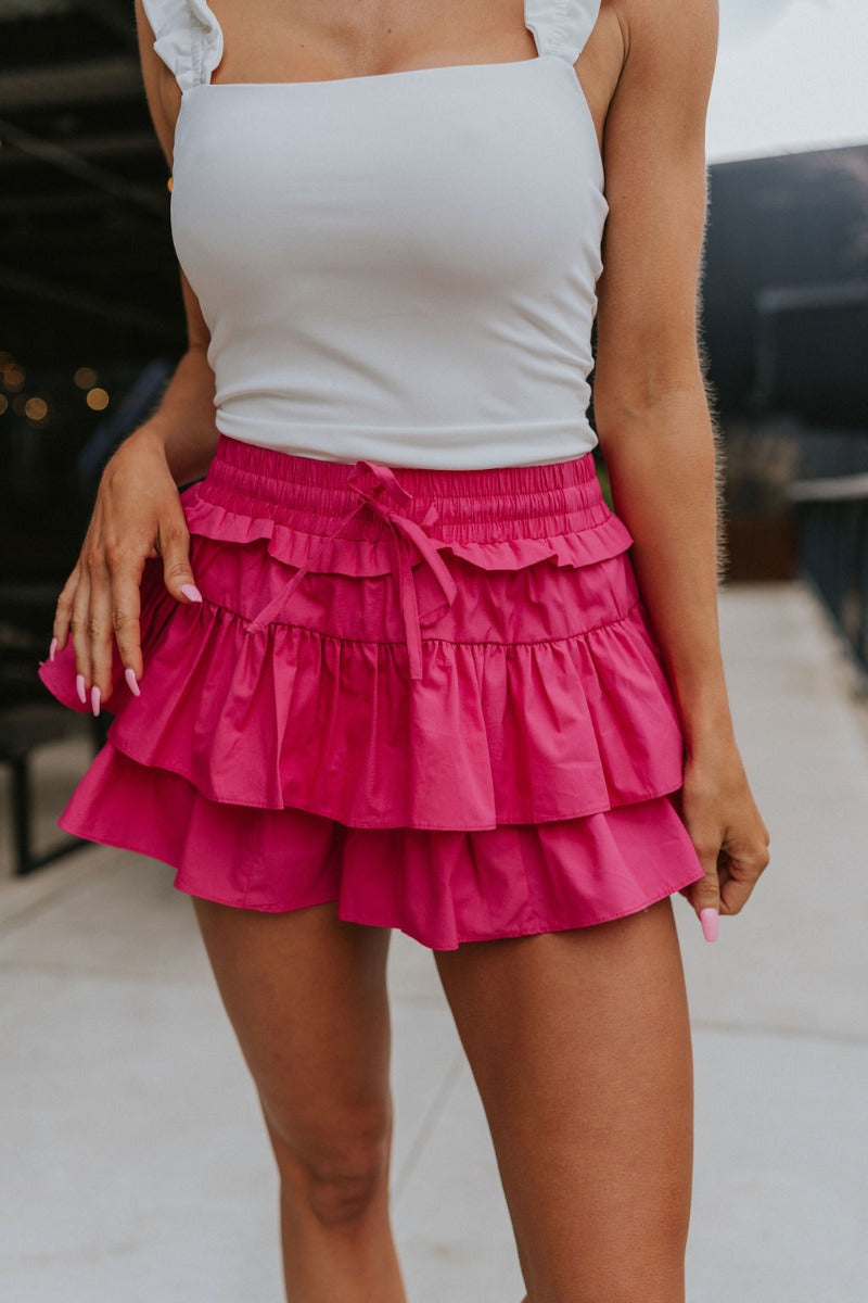 Front view of model wearing the Close To You Skort which features hot pink fabric, a skirt overlay with shorts underneath, a mini-length hem, ruffled tiered details, and an elastic/smocked waistband with a tie.