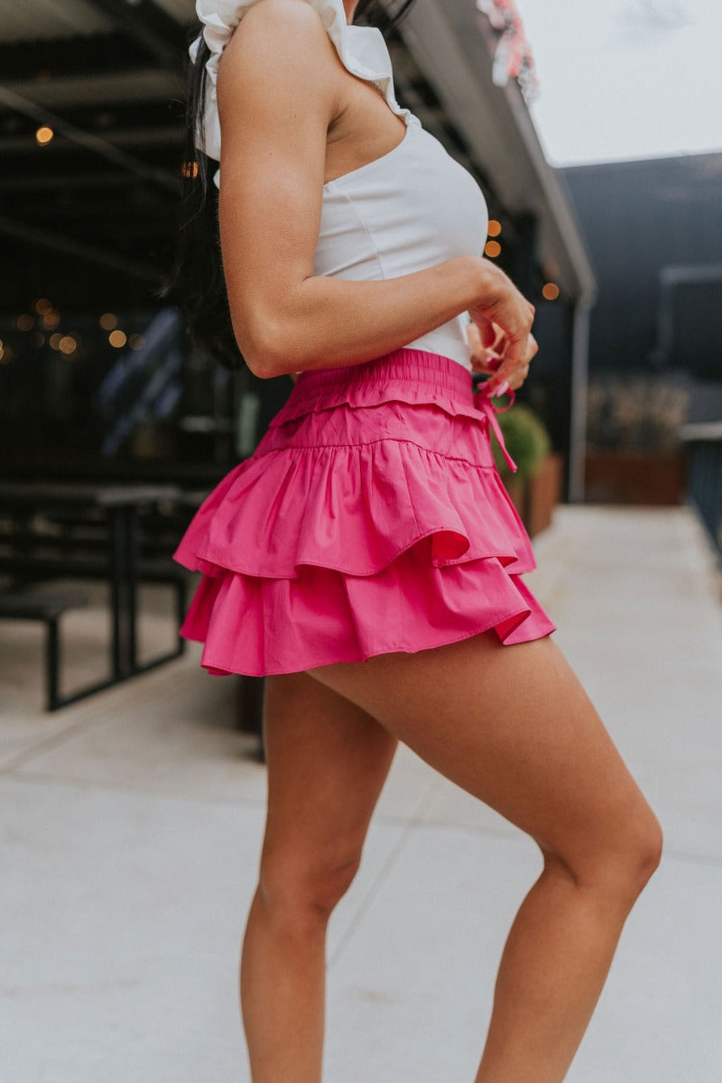 Side view of model wearing the Close To You Skort which features hot pink fabric, a skirt overlay with shorts underneath, a mini-length hem, ruffled tiered details, and an elastic/smocked waistband with a tie.
