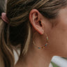 Close up view of model wearing the Rainbow Lake Earrings which features gold medium shaped hoops with multi color stones.