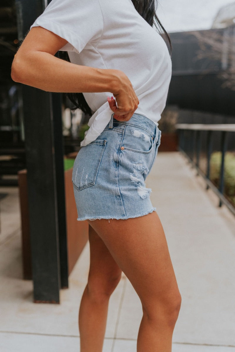Side view of model wearing the Just USA Denim: Sunny Day Shorts which features light-wash denim fabric, distressed detailing, two front pockets, two back pockets, a frayed hem, a front zipper with button closure, and belt loops on the waistband.