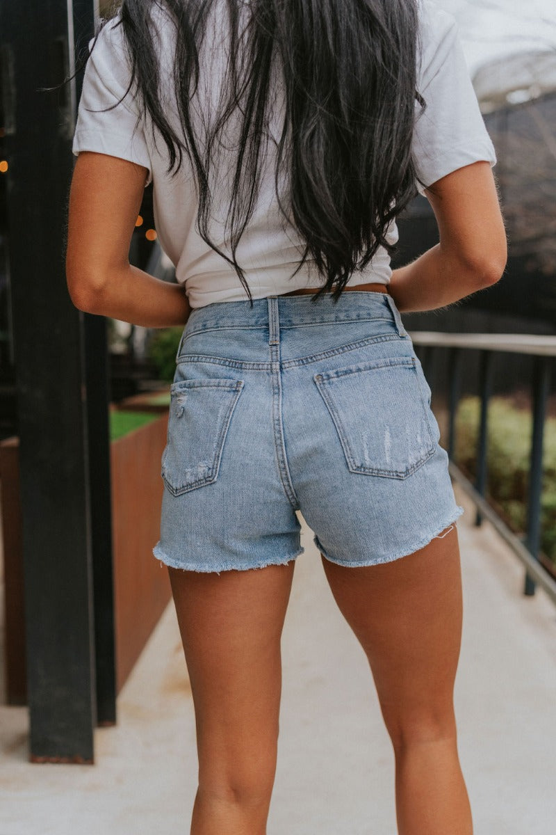 Back view of model wearing the Just USA Denim: Sunny Day Shorts which features light-wash denim fabric, distressed detailing, two front pockets, two back pockets, a frayed hem, a front zipper with button closure, and belt loops on the waistband.