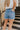 Back view of model wearing the Just USA Denim: Don't Think Twice Shorts which features medium-wash denim fabric, distressed detailing, two front pockets, two back pockets, a frayed hem, a front zipper with button closure, and belt loops on the waistband.