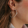 Close up view of model wearing the Reason To Be Earrings which features silver medium hoop shaped earring with roping details and clear stone attachment.
