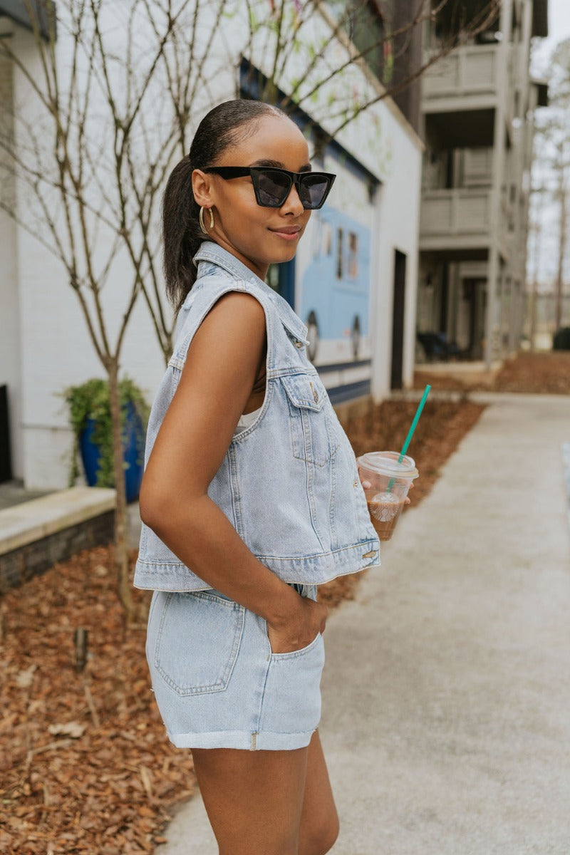 Side view of model wearing the Good For You Denim Vest that has light wash denim fabric, a cropped waist, silver buttons, a collar, front chest pockets, and distressed detailing, light brown stitching.