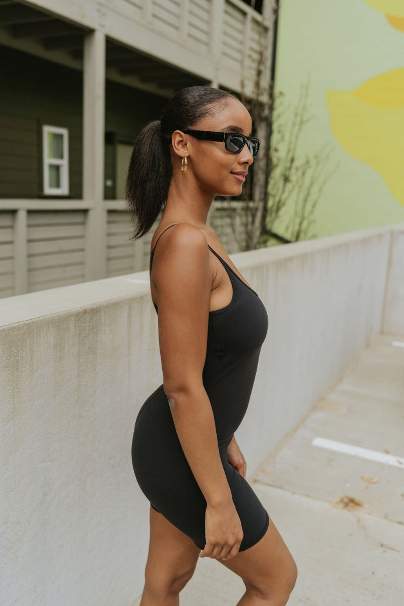 Side view of model wearing the Call It Even Romper that features black stretch fabric, a romper body, a round neckline, bra padding, and adjustable spaghetti straps.