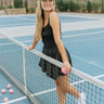 Full body front view of model wearing the Movement Mini Dress in Black that has a ribbed upper body, ribbed shorts lining, a lightweight skirt, a smocked waist, a scoop neck, and thick straps. Model is behind tennis net