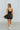 Full body back view of model wearing the Movement Mini Dress in Black that has a ribbed upper body, ribbed shorts lining, a lightweight skirt, a smocked waist, a scoop neck, and thick straps. 