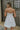 Back view of model wearing the Island Breeze Dress in White that has white fabric with a two-tiered baby doll style, a bubble hem, a plunge neck, adjustable straps, and an open back.