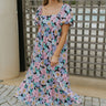 Full body front view of model wearing the Wild Blooms Midi Dress that has black fabric with a purple, pink, and blue floral pattern, ruffled hem, a square neck, a smocked back, and short puff sleeves.